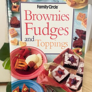 以下が含まれることがあります： Family Circle Brownies Fudges and Toppingsというタイトルの料理本。表紙には、イチゴがのった赤い皿やブラウニーが入った緑の箱など、カラフルな皿に盛られたブラウニー、ファッジ、デザートの写真が複数掲載されています。