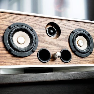 May include: A rectangular speaker with a white frame and a wood grain front panel. It features two large speakers with black rings, a smaller central speaker, and a silver knob. The speaker is designed for audio playback.