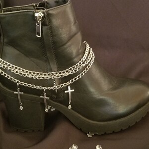 May include: A black ankle boot with a silver chain and cross charm ankle bracelet. The boot has a zipper closure and a chunky heel. The chain has three dangling crosses with small clear crystals. The boot is on a dark brown surface.