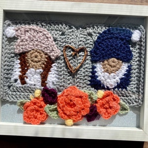 May include: A framed crocheted artwork featuring two gnomes with pink and blue hats, a heart-shaped twig, and orange and purple flowers.