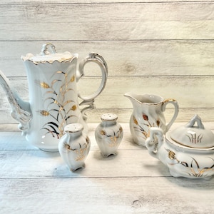 May include: A white porcelain tea set with gold accents. The set includes a teapot, creamer, sugar bowl, and two salt and pepper shakers. The design features gold wheat stalks and decorative handles.