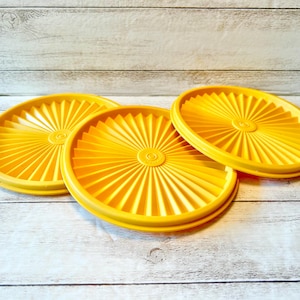 May include: Three bright yellow plastic lids with a radial design are arranged on a light wood-grain surface. The lids are circular with a raised center and a series of radiating lines, suggesting a vintage kitchenware set.