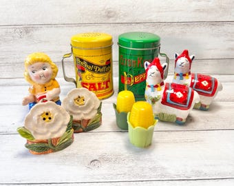 May include: A collection of decorative salt and pepper shakers and a salt tin. The shakers include a girl figurine, white flower, llama, and corn on the cob. The salt tin is yellow with black text that reads "Percival Duffin's SALT".