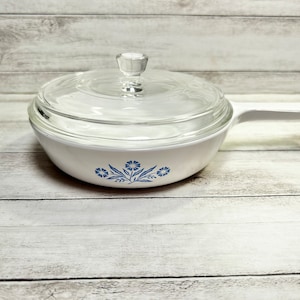 May include: White casserole dish with a blue floral design and a clear glass lid. The dish has a handle on one side.