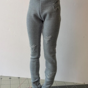 May include: Light gray doll-sized pants with distressed details. The pants have a textured fabric and feature ripped sections on the knees and thighs. The doll pants are designed to fit a small doll.