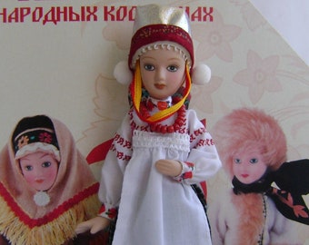 May include: A doll in traditional Russian folk costume, featuring a white embroidered blouse, a checkered skirt, and a white apron. The doll wears a headdress with red and gold accents. The background includes two other dolls and text in Russian.