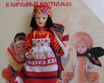 May include: Three dolls dressed in traditional Russian folk costumes. The doll in the center is wearing a red and white dress with a floral pattern and a colorful head scarf. The doll on the left is wearing a blue and white dress with a fur trim. The doll on the right is wearing a white fur coat with a black scarf. The dolls are all standing on a white background. The text "Куклы в народных костюмах" is written in red letters at the top of the image.
