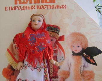 May include: Two dolls wearing traditional Russian folk costumes. The doll on the left is wearing a red and white dress with a red headscarf. The doll on the right is wearing a white fur coat with a brown fur hat. The dolls are standing on a white surface. The text "Куклы в народных костюмах" is visible on the background.