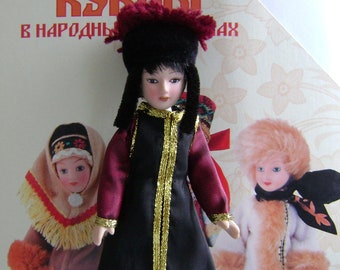 May include: A traditional Russian doll wearing a black dress with gold trim and a red hat with black fur. The doll is standing on a white surface. The text "Куклы в народных костюмах" is visible on the background.