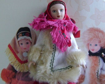 May include: Three Russian dolls wearing traditional folk costumes. The dolls are dressed in red, white, and pink with fur trim. The dolls are on a white background with the text "Куклы в народных костюмах" which translates to "Dolls in folk costumes".