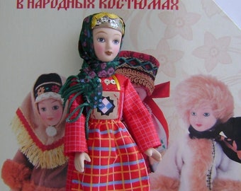 May include: A red, white, and blue plaid dress with a green scarf and a floral pattern. The dress has a black trim and a white and black geometric pattern at the bottom. The doll is wearing a silver crown and has brown hair and blue eyes. The doll is standing on a white background. The text "КУКЛЫ В НАРОДНЫХ КОСТЮМАХ" is visible on the background.