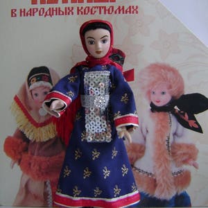 May include: A blue and gold dress with red trim and silver sequins. The doll is wearing a red headscarf and has brown hair and eyes. The doll is holding a red and white scarf in her right hand. The doll is standing on a white surface. The image is from a book titled "Куклы в народных костюмах".