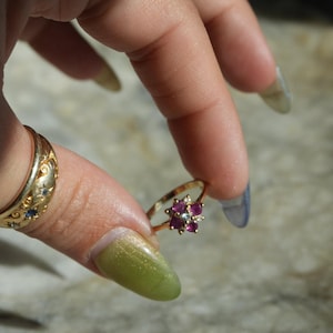 May include: A gold ring with a flower-shaped design, featuring red and white gemstones. The ring is held between fingers, showcasing its intricate details and craftsmanship. Another gold ring with blue gemstones is also visible.