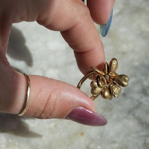 May include: A gold-colored ring with a flower design. The ring features a cluster of petals and a delicate band. The ring is held up by a hand with a gold band ring on the finger.