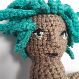May include: Crocheted doll with brown skin and teal hair. The doll has green eyes and a stitched-on mouth.
