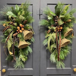 May include: A pair of matching teardrop-shaped door swags featuring green pine needles, magnolia leaves, and cedar sprigs. Each arrangement includes two woven tan bells, brown velvet ribbons, and clusters of small pinecones and brown nuts.