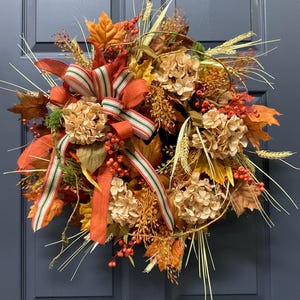 May include: A circular autumn wreath featuring a mix of orange, yellow, and brown maple leaves, dried beige hydrangea blossoms, red berry clusters, and tall wheat stalks. A decorative fabric bow with green, red, and white stripes is attached.