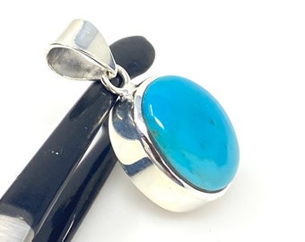 Puede incluir: Un colgante de plata con una piedra turquesa. La piedra es de forma ovalada y tiene un color azul claro.