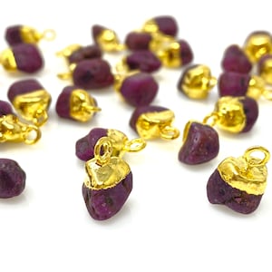 May include: A collection of raw, deep purple gemstone pendants with gold-colored metal caps and loops. The pendants are scattered against a white background, showcasing their natural, irregular shapes and metallic accents. These are ideal for jewelry making.