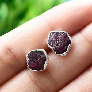 May include: A pair of silver earrings with raw ruby gemstones. The ruby stones are a deep red color and have a natural, irregular shape.