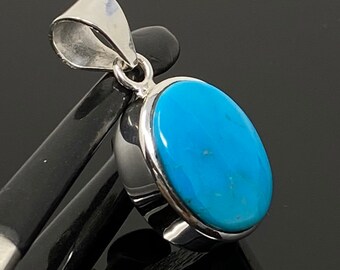 Puede incluir: Un colgante de plata con una piedra turquesa azul engastada en un engaste de bisel. El colgante tiene forma ovalada y tiene una simple anilla.
