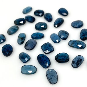 May include: A collection of 20 faceted, oval-shaped blue apatite gemstones. The stones are scattered on a white surface.