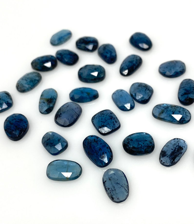 May include: A collection of 20 faceted, oval-shaped blue apatite gemstones. The stones are scattered on a white surface.