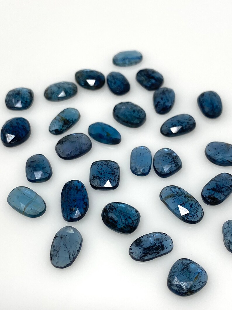 May include: A collection of 20 loose, faceted, blue-grey gemstone cabochons. The stones are oval shaped and have a speckled, cloudy appearance.