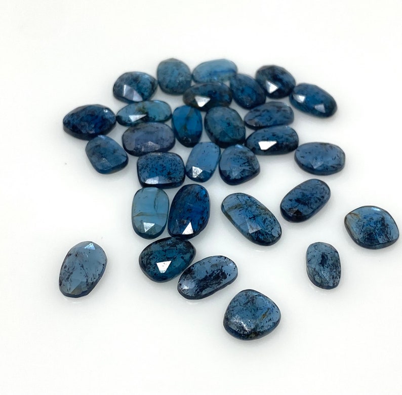 May include: A collection of 20 loose, faceted, oval-shaped blue kyanite gemstones. The stones are a deep blue color with a slight shimmer.