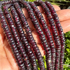 May include: A strand of dark red faceted gemstone beads. The beads are small and have a slightly irregular shape. The strand is long and is coiled in a hand.