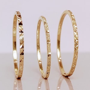 May include: Three gold stacking rings with different textures. The ring on the left has a diamond pattern, the middle ring has a row of small diamonds, and the ring on the right has a geometric pattern.