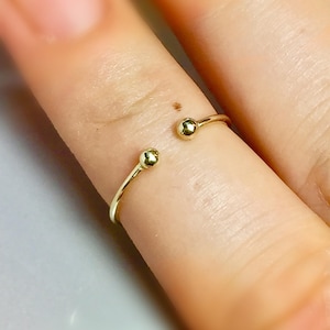 May include: A delicate gold-colored ring with a minimalist design. The ring features two small, spherical accents at the open ends. The ring is worn on a finger, showcasing its simple elegance. The ring's design is open, allowing for slight size adjustments.