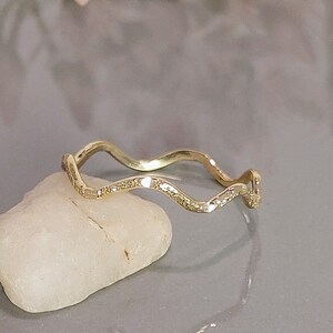 May include: A gold-colored, wavy ring with small, clear stones set along the top. The ring is displayed on a small, off-white stone against a gray background. The ring's design is a continuous, flowing wave.