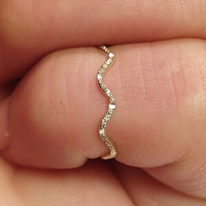 May include: A delicate, wavy gold ring with small, sparkling gemstones. The ring's design features a continuous, undulating pattern, creating a subtle yet eye-catching aesthetic. The gemstones are evenly spaced along the wave, adding a touch of elegance.