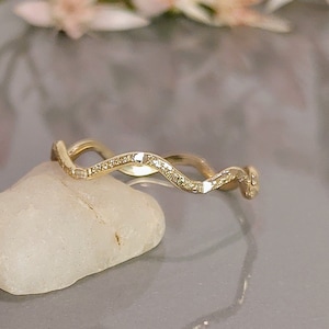 May include: A gold-colored ring with a wavy design, set with small, sparkling stones. The ring is displayed on a white stone against a soft, blurred background. The ring's design is elegant and eye-catching.