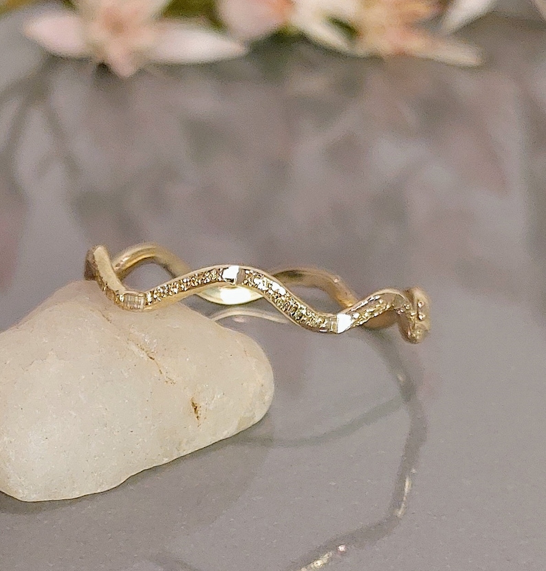 May include: A gold-colored ring with a wavy design, set with small, sparkling stones. The ring is displayed on a white stone against a soft, blurred background. The ring's design is elegant and eye-catching.