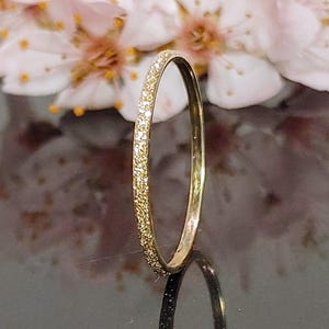 May include: A thin gold band ring with a textured surface. The ring is on a black surface with a blurred background of pink flowers.