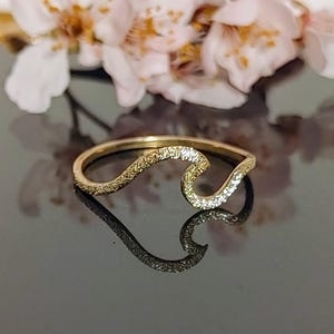 May include: A gold-colored ring in a wave design, with a textured surface on one side and small, sparkling stones on the other. The ring is set against a dark, reflective surface, with soft pink and white flowers in the background.