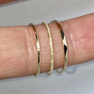 May include: Three gold stacking rings. The ring on the left is a plain band. The middle ring is a thin band with small diamonds. The ring on the right is a hammered band.