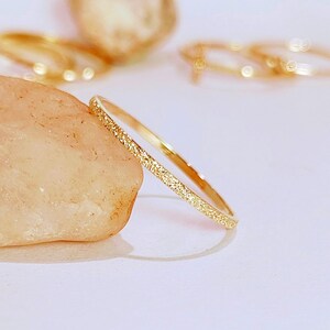 May include: A thin gold band ring with a textured surface. The ring is made of metal and has a simple design.