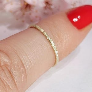 May include: A delicate gold ring with small, sparkling stones set along the band. The ring is shown on a finger with red nail polish, with soft pink flowers in the background. The ring appears to be a wedding band or a stacking ring.