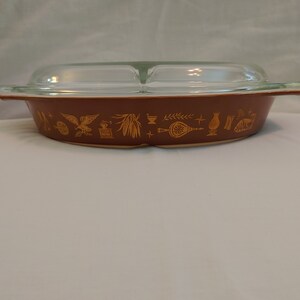 May include: A brown oval casserole dish with a clear glass lid. The dish has a gold trim with a pattern of various American symbols, including an eagle, a wheat stalk, a liberty bell, and a cornucopia.