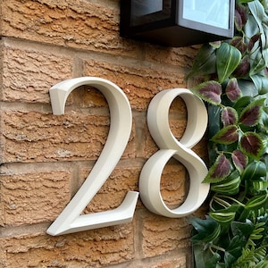 May include: Cream-coloured house numbers "28" fixed to a brick wall. The numbers are large, three-dimensional, and have a modern design. The wall is made of red-brown bricks. A black light fixture and greenery are visible in the background.