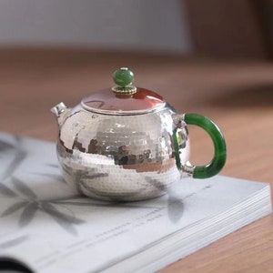 Op de afbeelding: Een zilveren theepot met een gehamerde afwerking en een groene jadeknop op de deksel. De theepot heeft een groene jadehendel en staat op een boek.