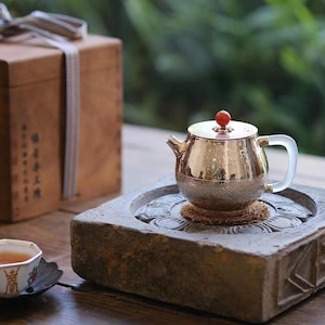 Może przedstawiać: Srebrny czajnik z czerwonym pokrętłem na pokrywce stoi na kamiennej podstawie z rzeźbionym wzorem. Mały kubek do herbaty z kwiatowym wzorem stoi na czarnym spodeczku obok czajnika.