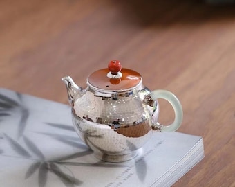 Puede incluir: Una tetera de plata con un acabado martillado y un pomo rojo en la tapa. La tetera tiene un asa de jade verde y está sentada sobre una pila de libros.