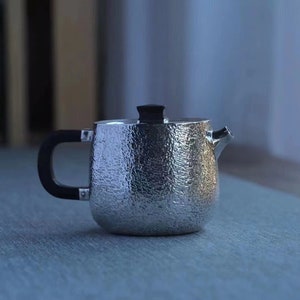 May include: A silver teapot with a hammered texture and a black handle. The teapot is sitting on a blue surface.