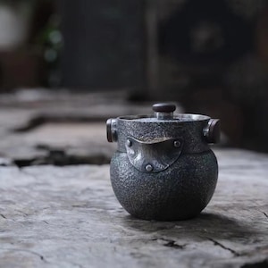 Puede incluir: Una tetera de plata con una superficie texturizada y una perilla de madera en la tapa. La tetera tiene dos pequeñas asas a los lados y una pieza de metal decorativa en la parte delantera.