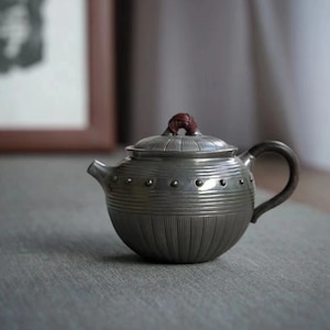 Pode incluir: Uma chaleira de prata com uma superfície texturizada e nervurada e um botão castanho na tampa. A chaleira tem um corpo redondo e um bico curvo.