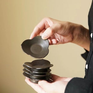 Puede incluir: Un conjunto de cinco platillos para tazas de té de cerámica negra, cada uno con un borde festoneado, se sostiene en las manos de una persona. El platillo superior se está levantando de la pila.